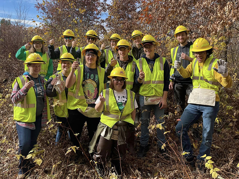 Natural Resources Club happy, thumbs up group photo in the woods with yellow hard hats and reflective vests