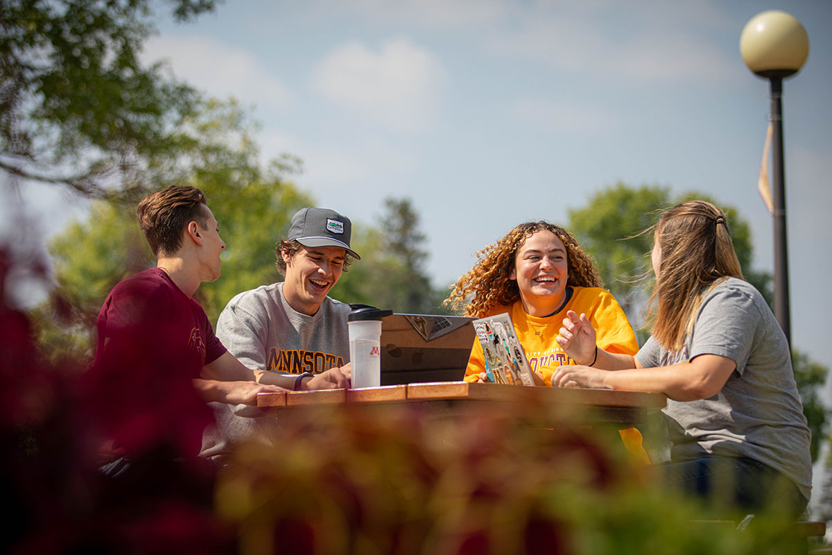 Students laughing while having a conversation at a picnic table on campus.