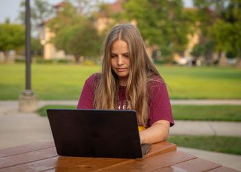 Female student typing on her laptop outside near the Campus Mall at a picnic table