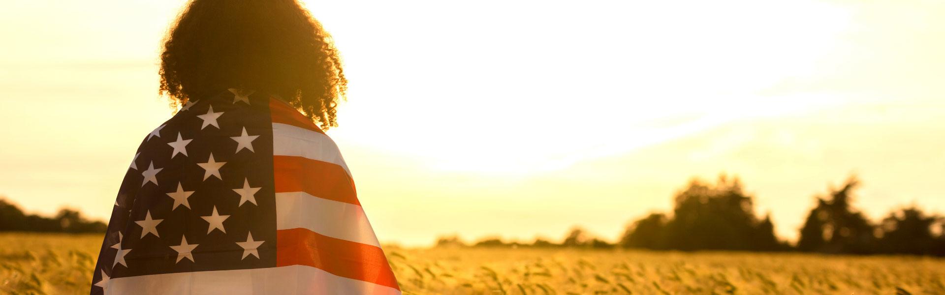 Woman wrapped in an American Flag standing in a wheat field at sunset