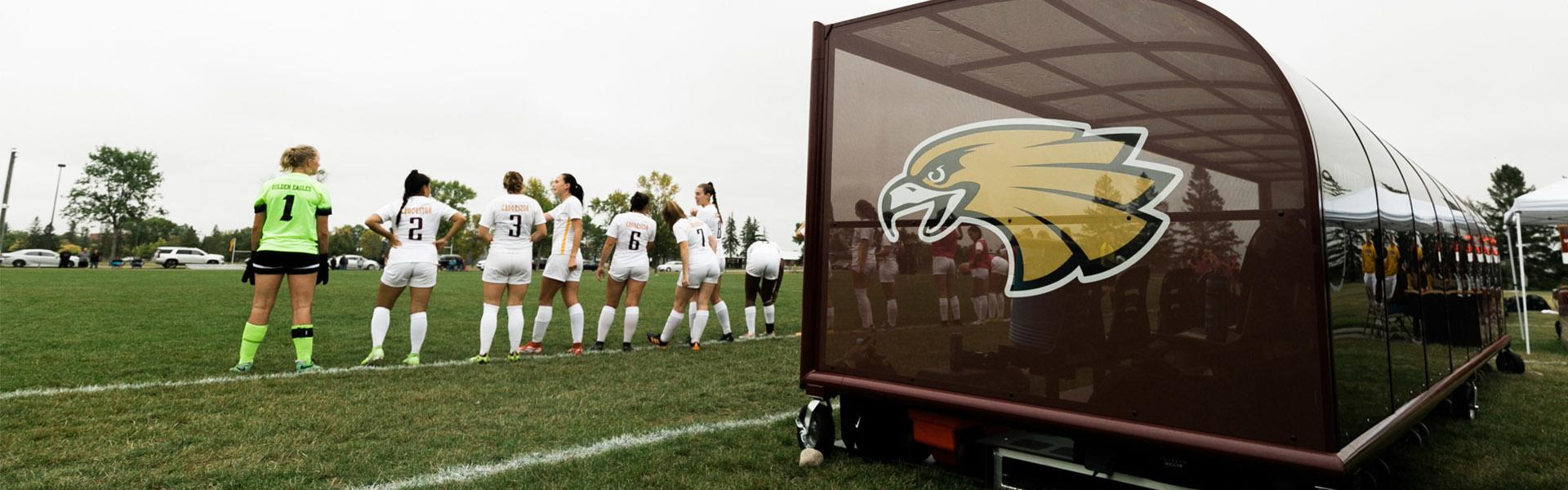 Women's soccer players standing on the sideline with the Golden Eagle logo on the dugout