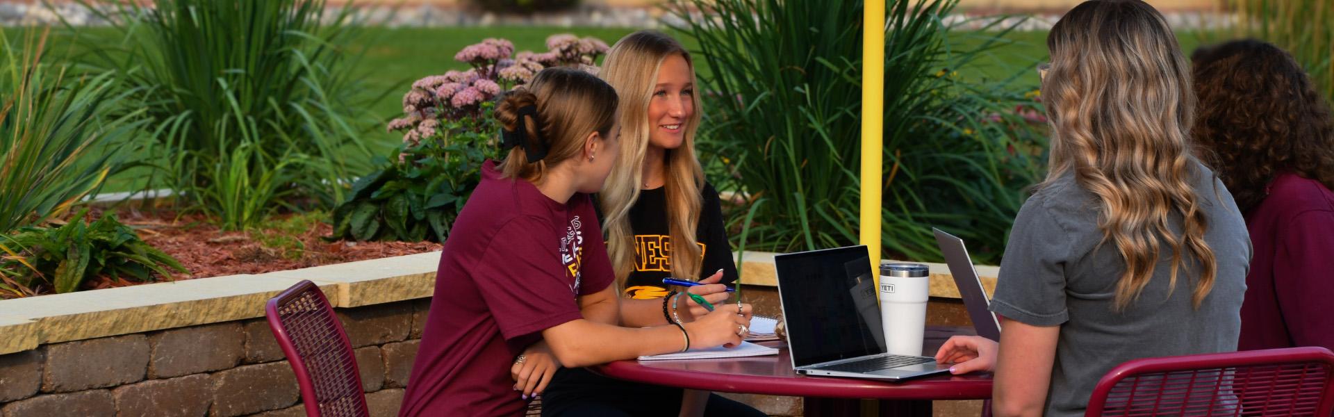 Four female students with notebooks and laptops sitting at a maroon and gold picnic table in the summer chatting.