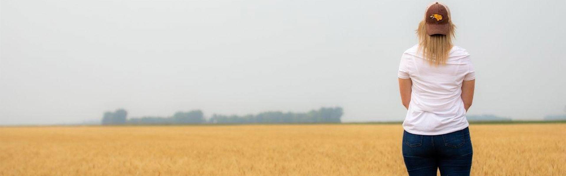 Blonde hair female standing in a wheat field with her back to the camera wearing a Golden Eagle hat