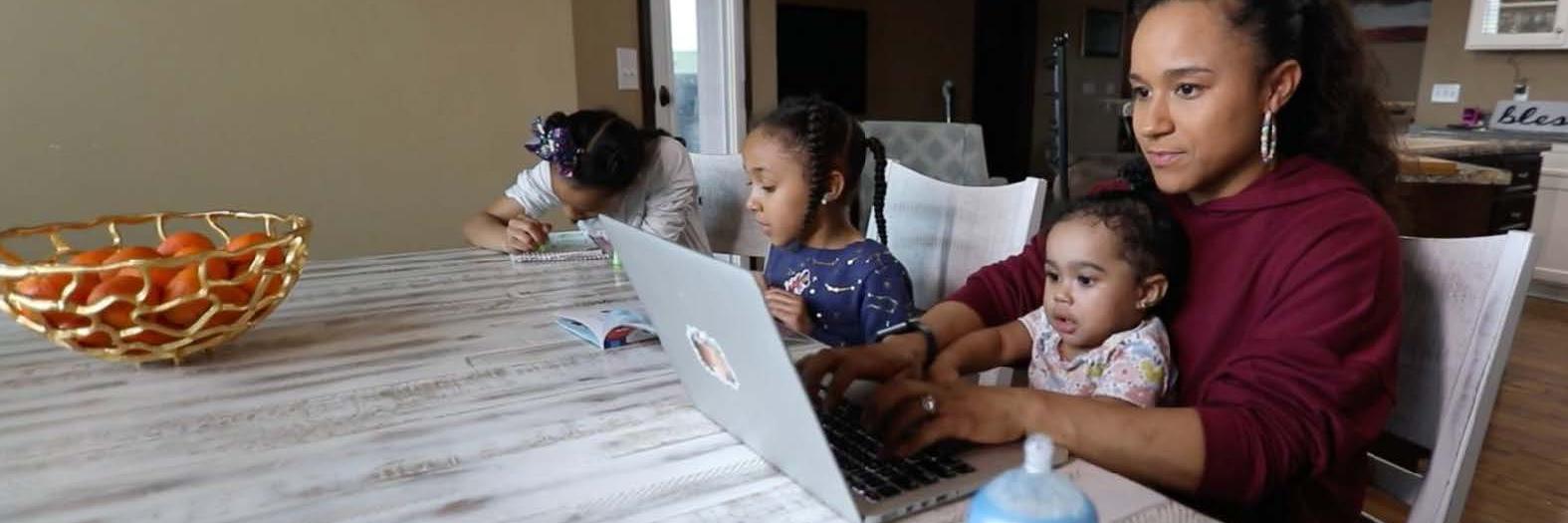 A student working on their laptop at a table with two children nearby and one seated on their lap.