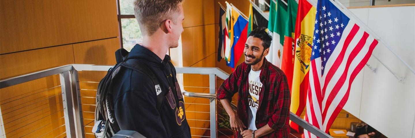 Two students in the student center talking on the steps to second floor with several international flags behind them.