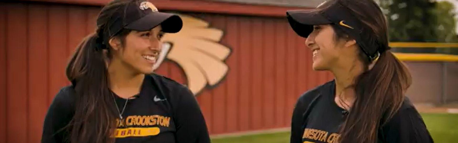 Leah and Hannah Marcias in their softball gear laughing and smiling while giving an interview near the softball dugout