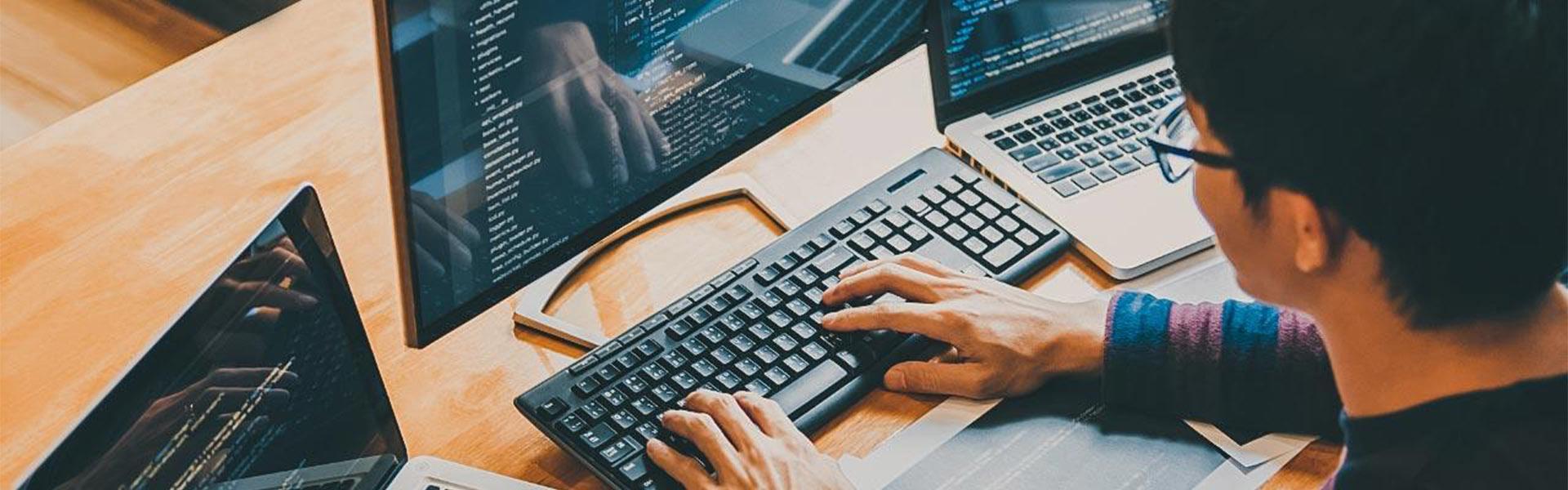 Male sitting at a desk on a keyboard with a few monitors and a laptop, all with programming languages scrolling across the screens