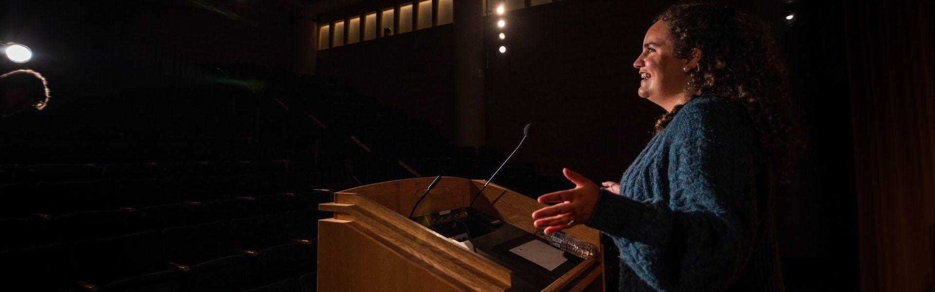 Woman standing at podium in a large theatre speaking to the crowd 