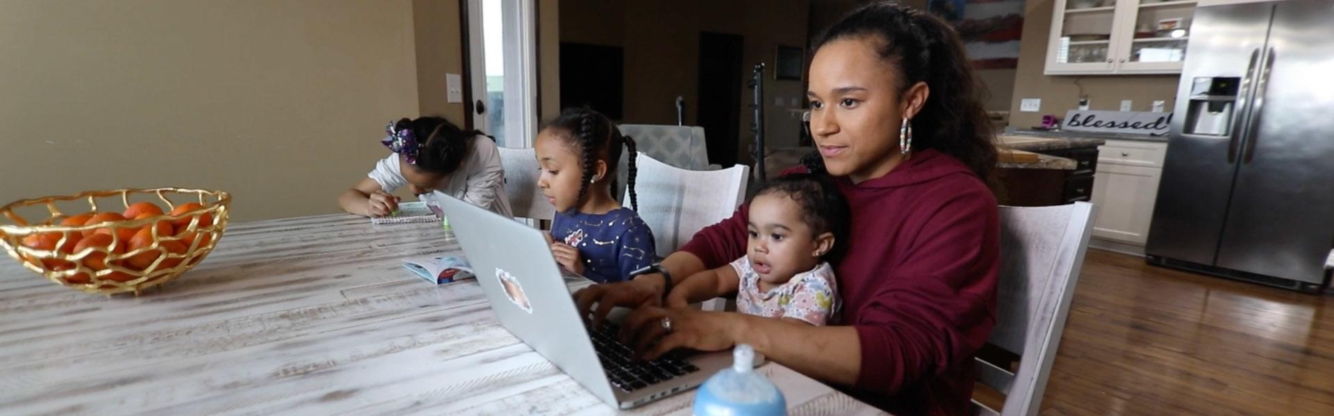 A mom of three young kids, one on her lap, while she's working on a laptop at the kitchen table