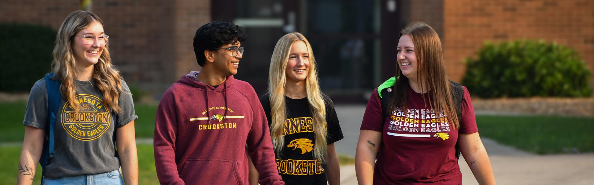 Group of four diverse students walking outside of Hill Hall and Sargeant Student Center in early fall talking and smiling