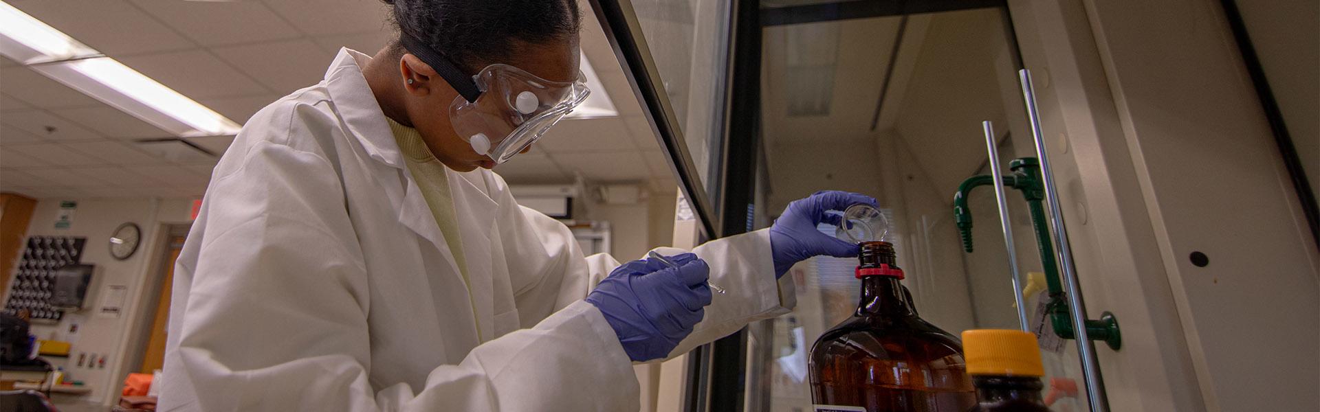 Student with a lab coat, safety goggles and blue gloves in the Dowell Hall Lab pouring chemicals into a container