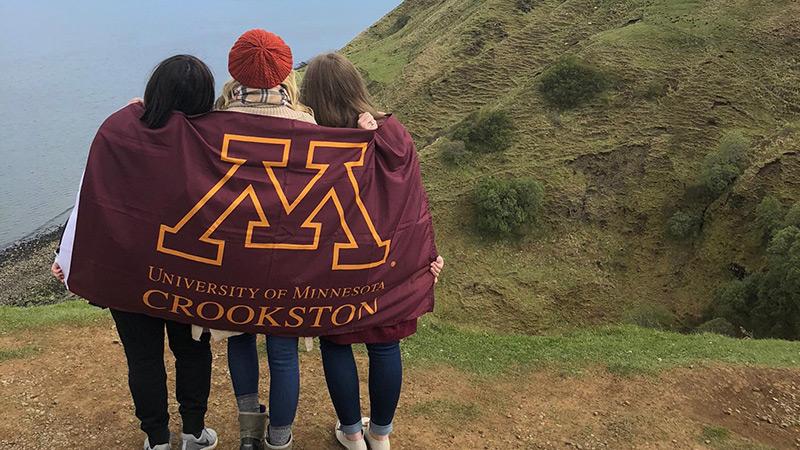Three students wrapped in a UMN Crookston flag a top a hill overlooking the ocean