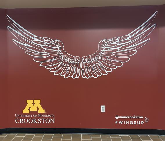 Life size white painted eagle wings on a mroon background and the M Crookston logo and painted #wingsup hashtag near the Eagles Nest