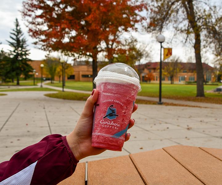 Person holding a Caribou pink refresher outside Sargeant Student Center with beautiful fall colors in the background