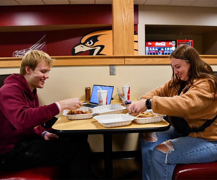 A male and female students sitting in a booth outside Regal's grill with their burgers and sandwiches, fries and laptops talking