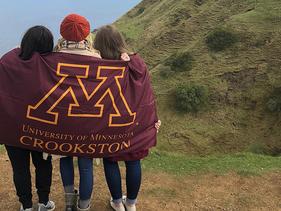 Three female students standing with a University of Minnesota Crookston flag wrapped around them on a hill looking out on the ocean on a Study Abroad trip