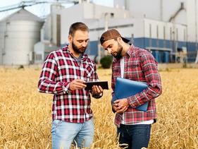 Two men in a grainfield looking at a tablet with a grain elevator in the background