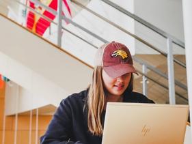 Female student sitting in the Northern Lights Lounge with a Golden Eagle hat on a laptop