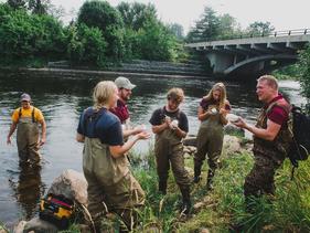 Faculty and students on a riverbank chatting and taking water samples