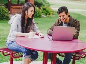 Two international students sitting outside talking and smiling looking at a laptop